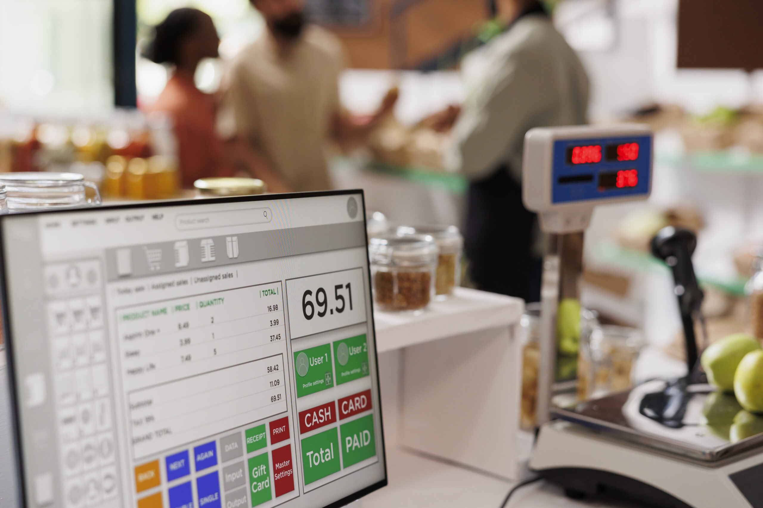 A Point of Sale (POS) monitor displaying an inventory and sales interface at a retail checkout counter in Malaysia, with a digital scale and customers in the background.